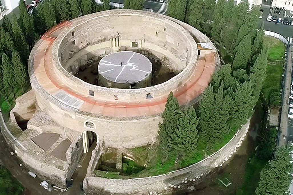 An aerial shot of a large circular building, with reddish and yellow stones, surrounded by trees on all sides