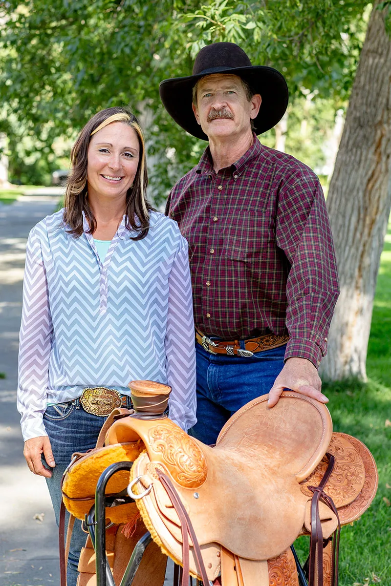 A woman and man pose behind a brown leather saddle outdoors.