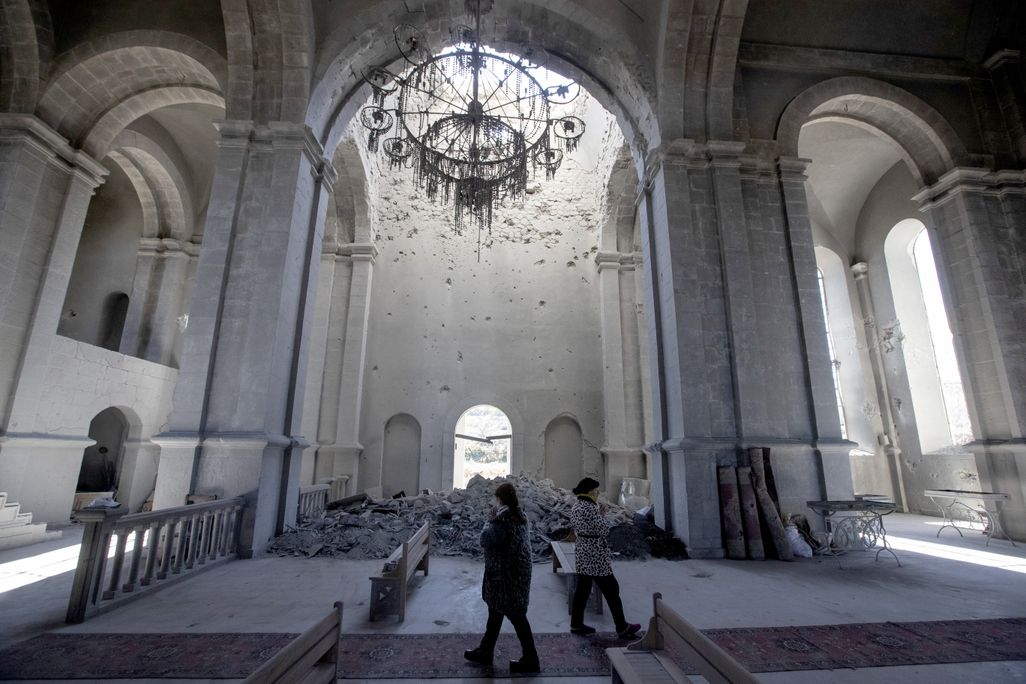 Two people walk, cast in shadows, underneath the tall white arches of a cathedral; behind them, a pile of rubble blocks a doorway and a broken chandelier hangs from the ceiling