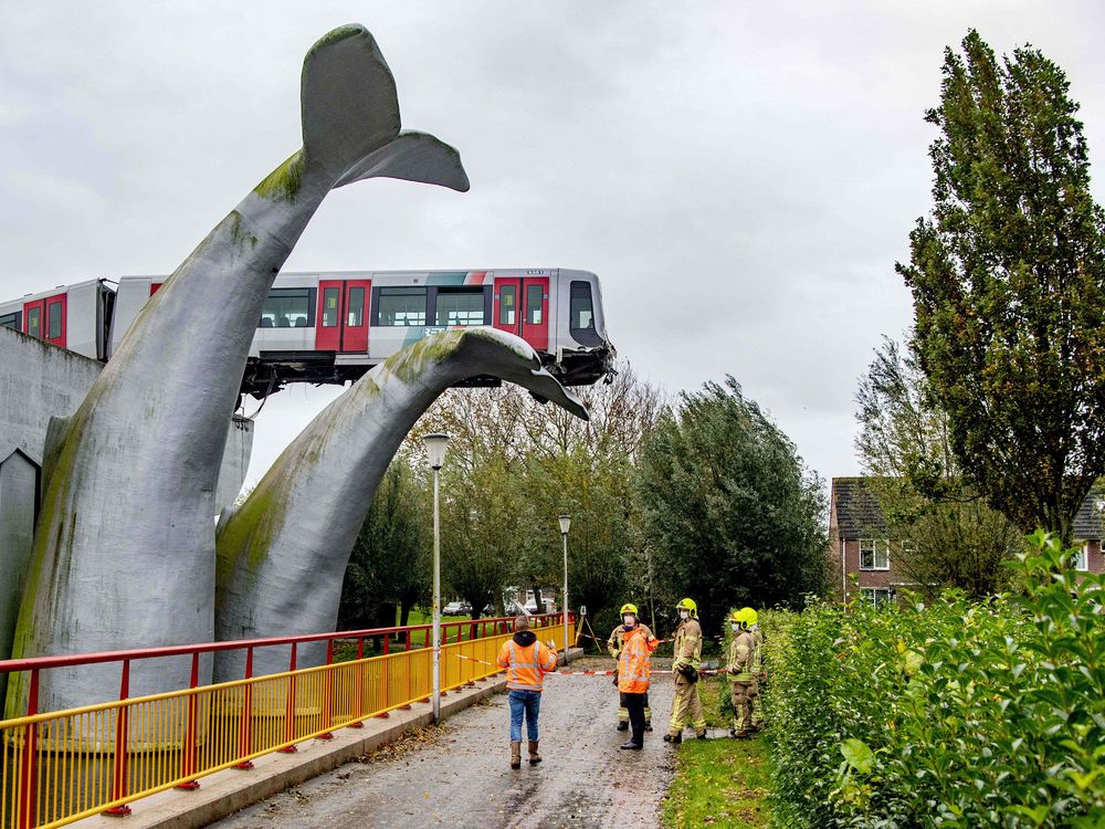 A red and silver metro train rests precariously, hanging off of the train tracks, with the first car resting on one of two enormous sculptures of whale tails. Below, six construction workers asses the damage. 