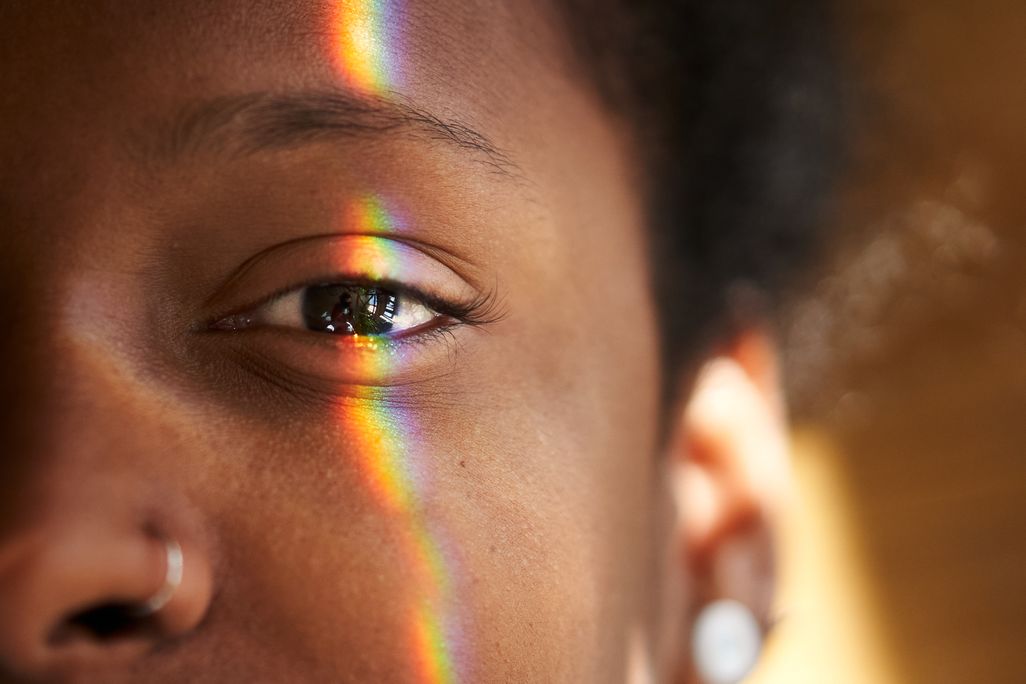 half of a Black woman's face as she looks into the camera with a rainbow strip of light across her eye