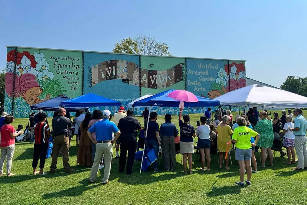 A crowd gathering under and around shade tents set up in front of the mural under a blue sky.