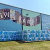 Turquoise mural covering the entire side of a building, with a pattern of waves along the bottom, and a brown textile with the Spanish words Viva la Vida! in the center.