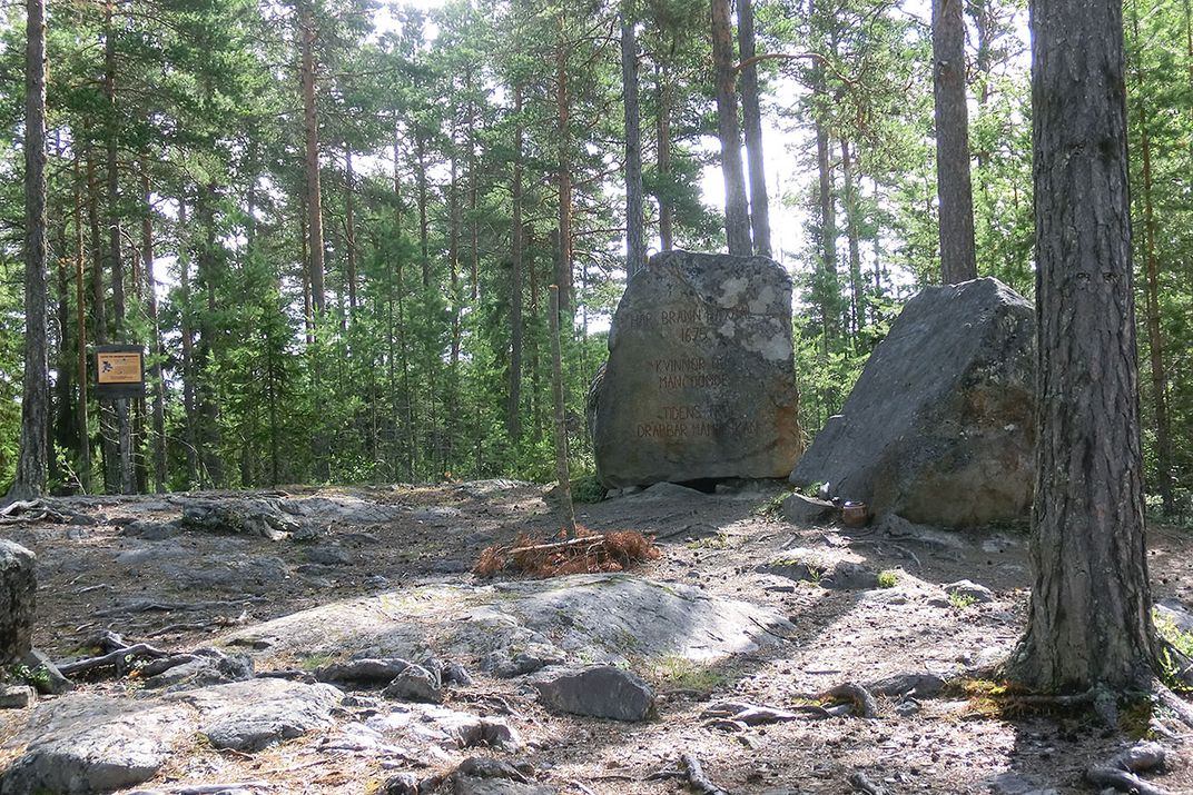 A clearing in the woods, with two large boulders carved with names. At the far end of a clearing, a sign is posted to a tree, but it is too small to read.