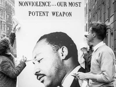 Two civilians look at a poster featuring the face of Martin Luther King Jr., his head depicted beneath the words 'Nonviolence... Our Most Potent Weapon.'