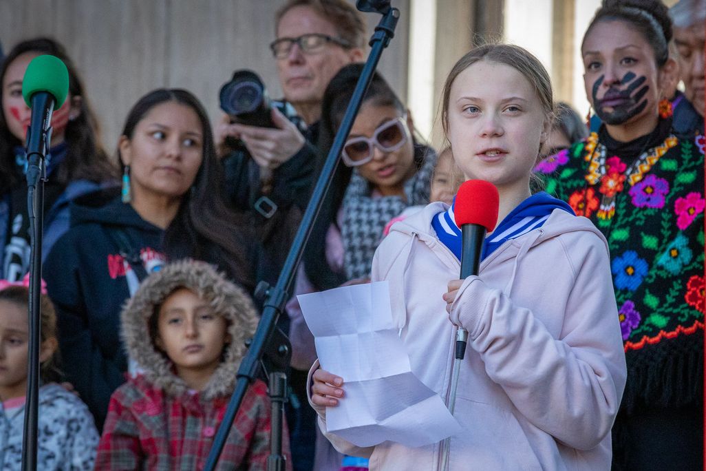 young blond girl in white coat with blue color holding a red microphone with speech in hand speaking in front of young kids