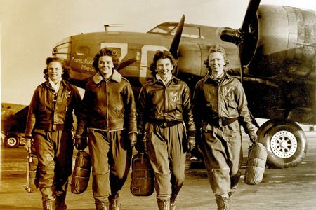 A black-and-white photo of four white women in their late 20s to early 30s walking on a military airfield. The women are wearing flightsuits, boots, and leather flight jackets. Behind the women is the forward fuselage of a World War II military bomber.