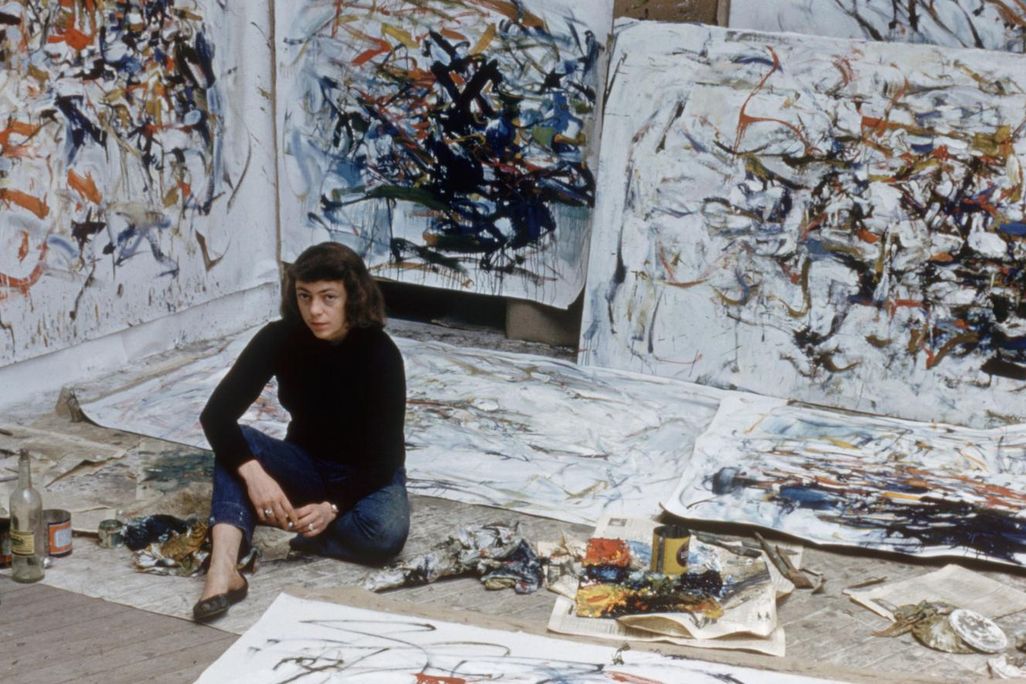 A view of Mitchell, wearing a black turtleneck, seated amid an explosion of white canvases with markings