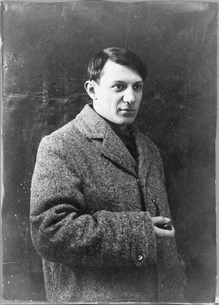 A black and white photograph of a young man, Picasso, standing and wearing a thick gray coat, holding a pipe and staring into the distance to the viewer's right