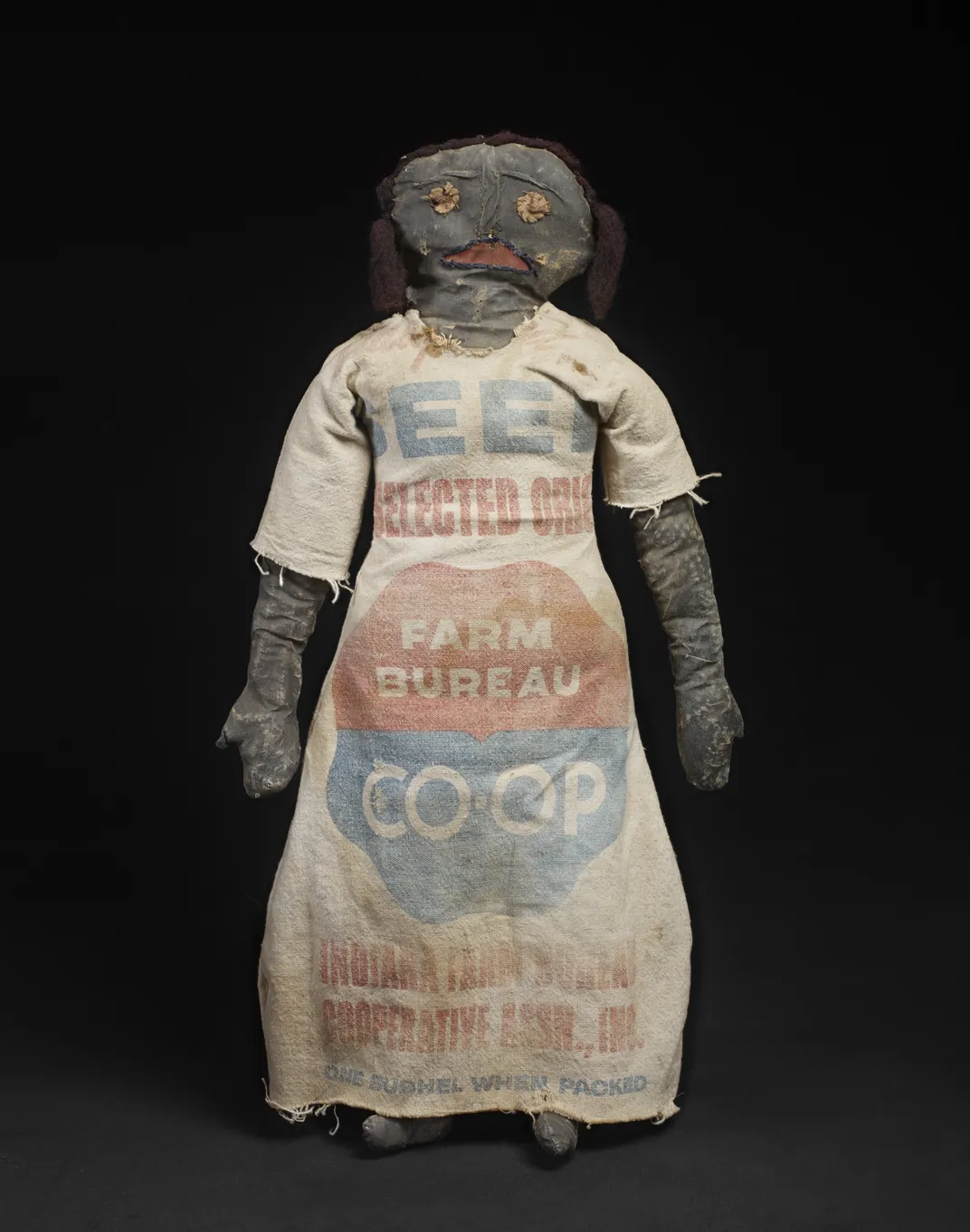 A female Black doll, worn by time, wearing a dress made out of a recycled feed sack
