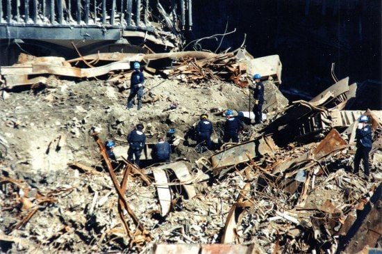  Photo of the destruction after September 11, 2001, in New York City. Workers in blue helmets and uniforms scramble around a pile of destruction, bent metal, and ruins.