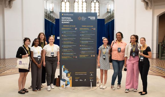A group of teachers gather around a large blue billboard of the Summit schedule