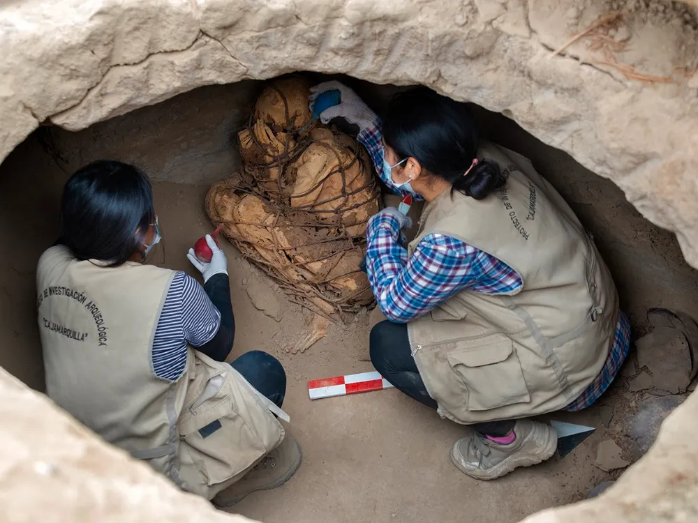 Two people kneel in an oval-sized opening and carefully brush the mummified body