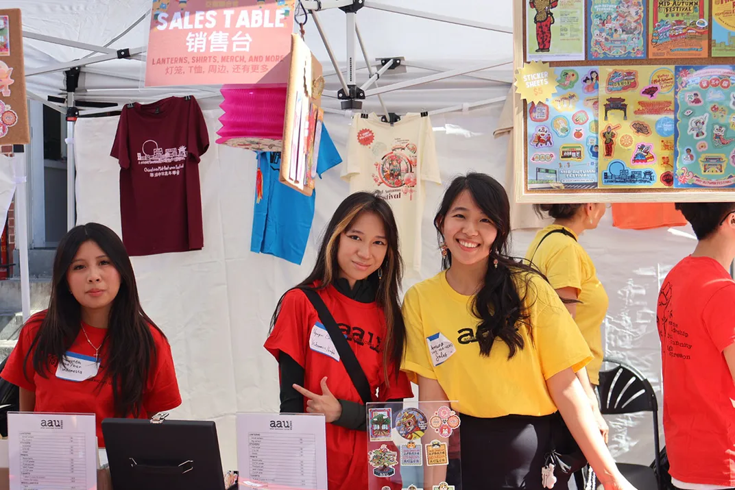 Three young adults with long dark hair, wearing matching red or yellow T-shirts, pose from behind the counter of an information booth.