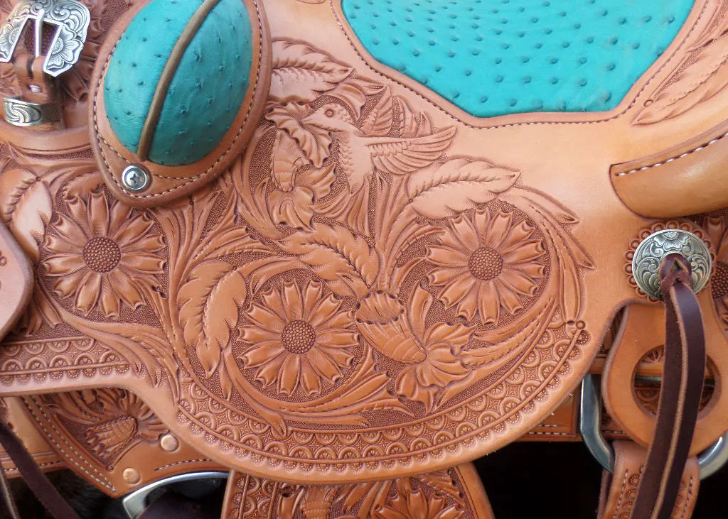 Close-up on the side of a brown leather saddle with turquoise accents. The brown leather contained etched designs including floral patterns and a hummingbird drinking from a flower.
