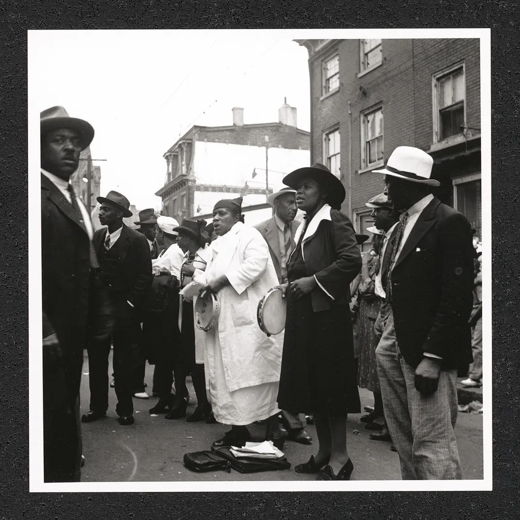 A woman sings, holding a tambourine, surrounded by men in suits outdoors. Black-and-white photo.