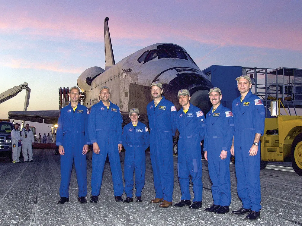 Seven people wearing blue flightsuits stand in front of a space shuttle parked outside.