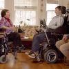 Film still of an elder woman and a young man, both in wheelchairs, facing each other in conversation and smiling in a warmly lit living room. A man sits to the side, next to a laptop, also smiling.