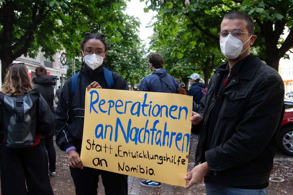 Two people in masks hold a yellow sign with blue and black handpainted letters, standing in a city street in front of trees and other protesters