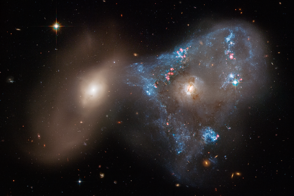 An image taken by the Hubble Space Telescope of two galaxies interacting with eachother. The galaxy on the right is surrounded by sparkling blue young stars. The galaxy towards the left is smaller and is not as flashy.