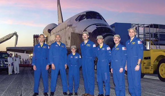 Seven people wearing blue flightsuits stand in front of a space shuttle parked outside.