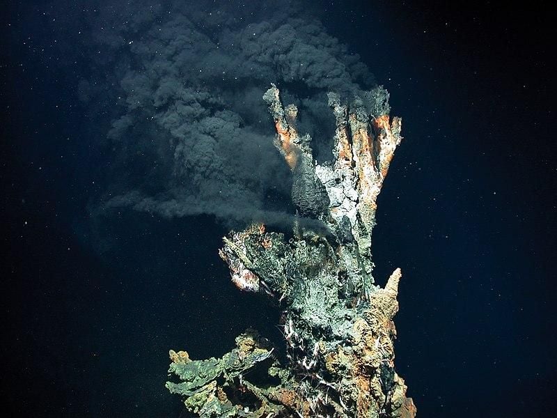 A hydrothermal vent in the deep ocean produces a dark, cloud of hot air against a dark blue background. The vent in the foreground is rocky, like a conglomerate of shells and rocks piled on top of each other.  