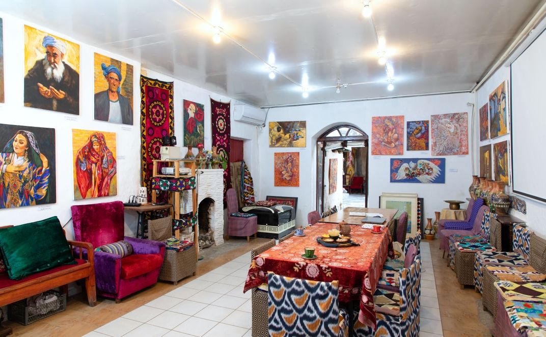 A room in Human House is filled with tables, chairs, and crafts. In the middle of the room, two rectangular tables are pushed together. Along the walls, several paintings and carpets are hung. Chairs line the perimeter of the room against the walls.