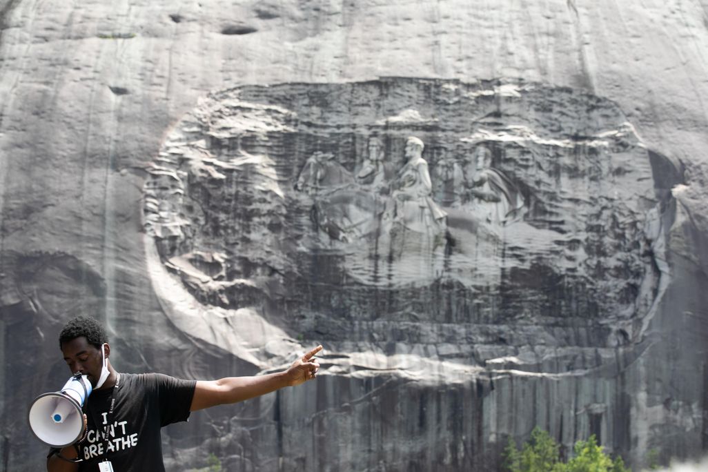 A Black man wears a black and white t shirt and stands with a megaphone, gesturing to the Confederate monument on the face of a gray granite rock behind him