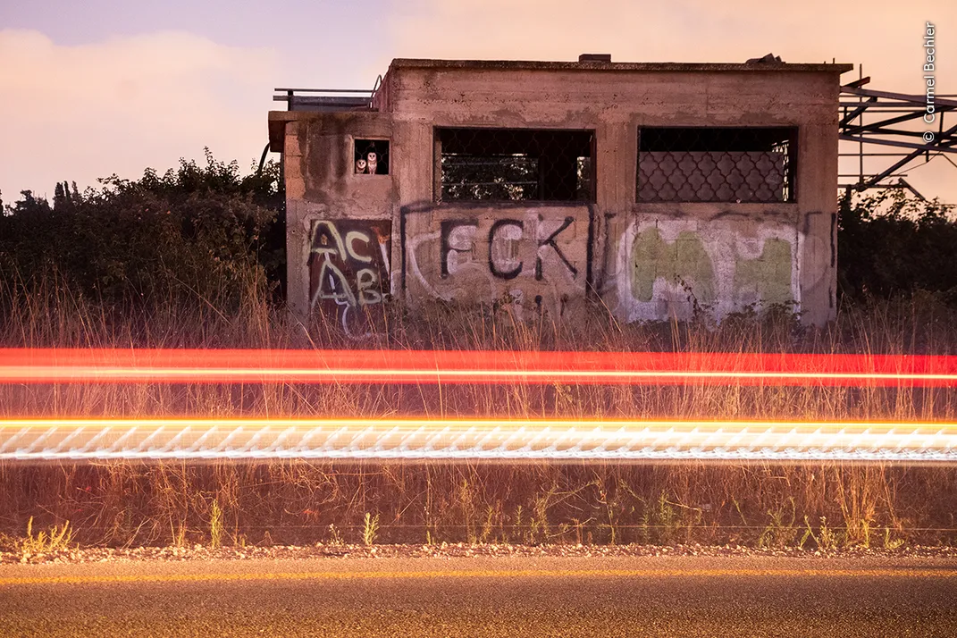 an abandoned building on the side of a road; two streaks of light cross horizontally in the middle of the image; two white owls look out the smallest window of the building
