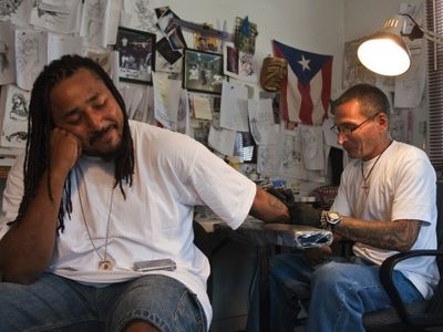 Q Monts sits patiently at Nu Flava Ink while tattoo artist Charles “Coco” Bayron puts the finishing touches on Q’s tattoo reading “Time is Money, Money is Time.”