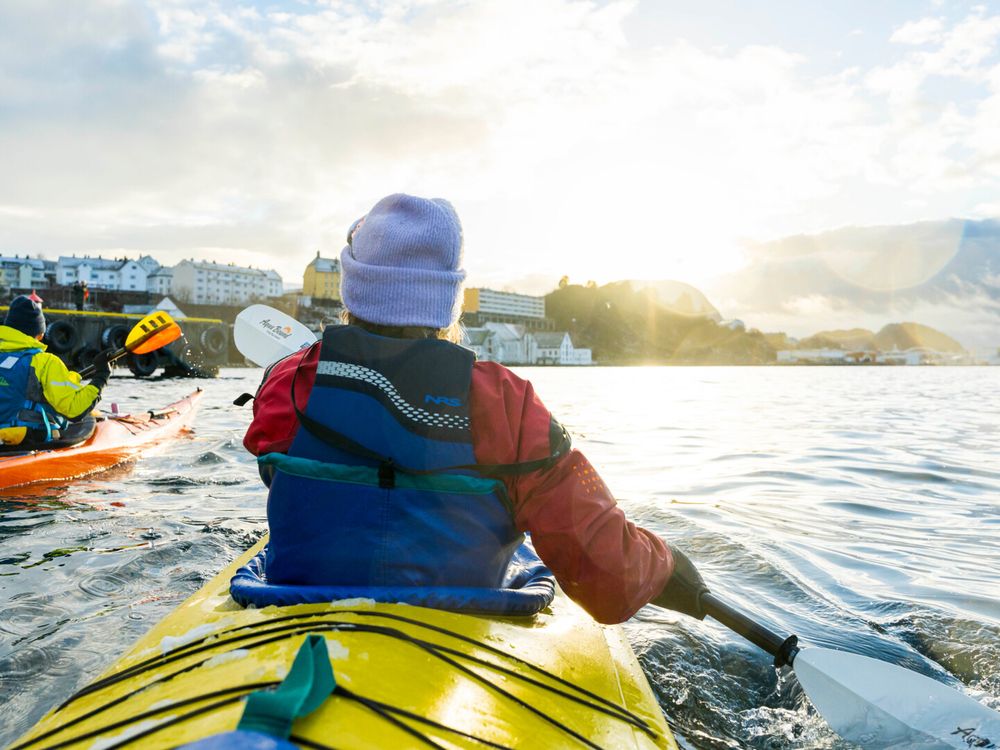 Two kayakers paddle through Ålesund’s waters with Uteguiden, surrounded by shoreline buildings and hills under a partly cloudy sky.