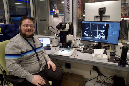 A man in a grey short sits in a lab setting, in front of a microscope and a computer screen.