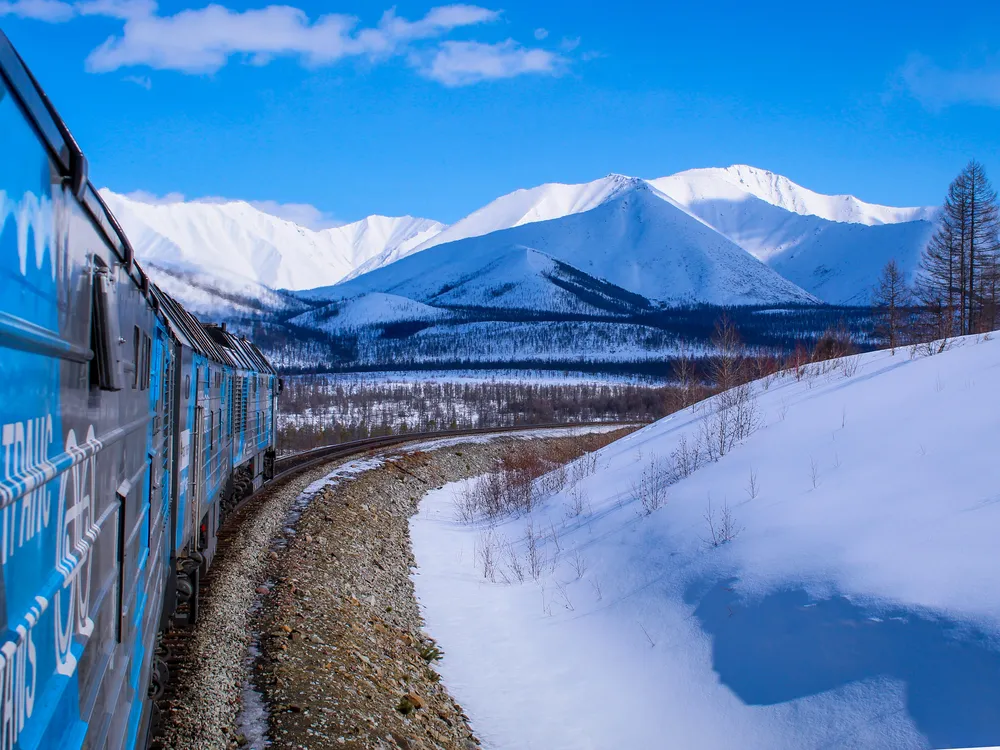 13 - A train whizzes through snowy hills with frosty peaks from the Stanovoy Ridge mountains in the distance.