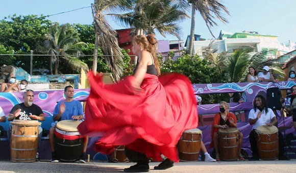 A woman in a long, flowing red skirt, blurry with motion, dances in front of a line of people seated and playing barrel-shaped drums. Palm trees sway in the background.