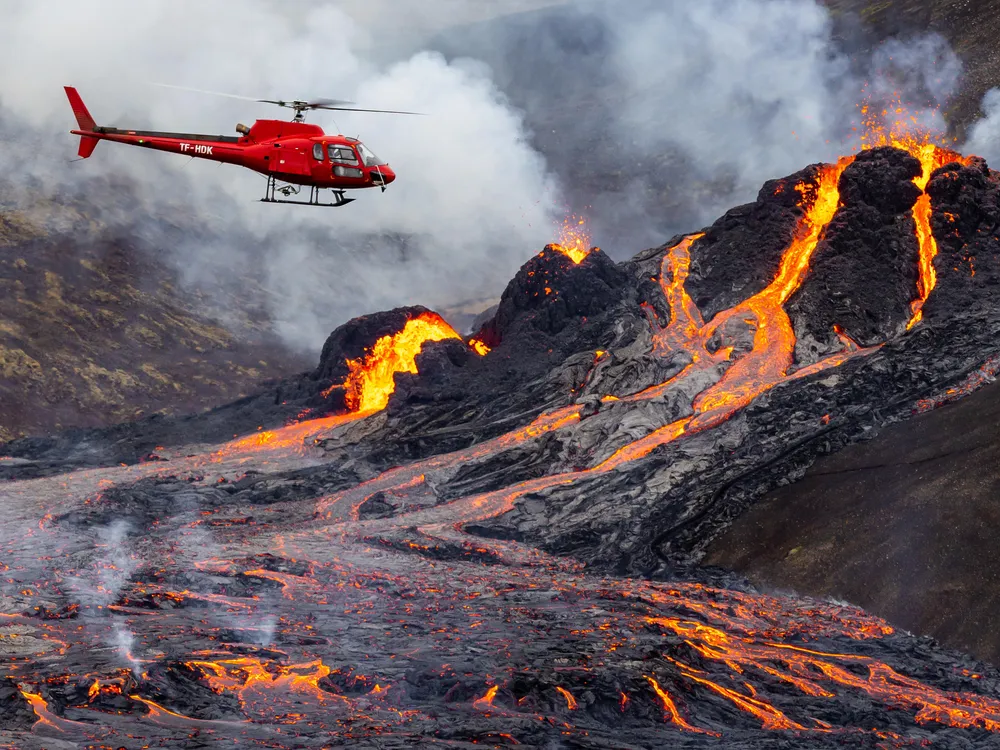 The photo shows a volcanic eruption. Magma is flowing down the volcano. A red helicopter hovers near the volcano.