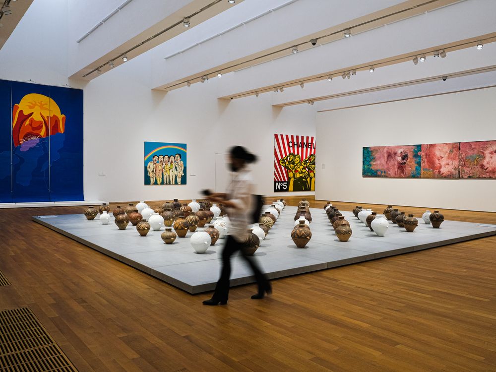 An interior view of a gallery in museum, with tall white ceilings, wooden floor and in the center, a work of 126 clay jars, some painted white, arranged on the floor in a grid