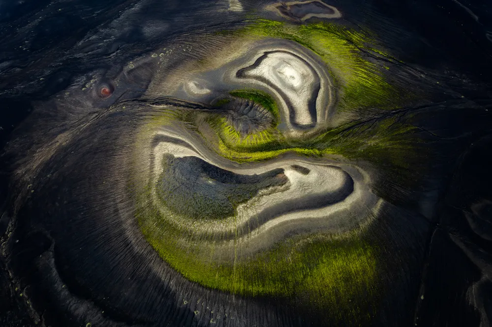 This abstract aerial photo captures the Icelandic highlands from above. A black volcanic hill covered in green moss surrounds the center while layers of black and white shades create intriguing patterns. In the top left corner, a small red crater adds a pop of color to the scene.