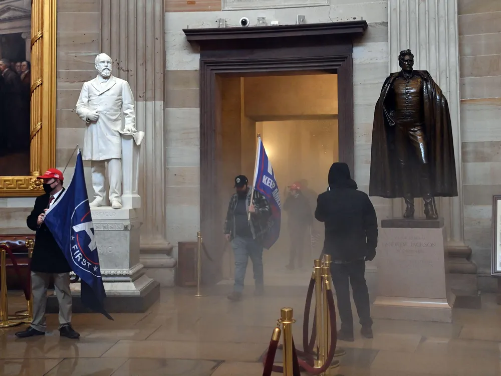 Two men carrying large blue flags - the first words of KEEP AMERICA GREAT appear in one - walk into the corridor flanked by two statues of men and huge Trumbull paintings in gilded frames, surrounded by hazy gas on the floor