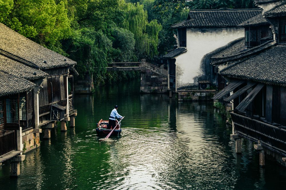 In Wuzhen, China, in the afternoon, a boatman moves slowly down a calm river. This peaceful scene, surrounded by old wooden buildings and lots of greenery, shows how beautiful and peaceful this ancient water town has always been.