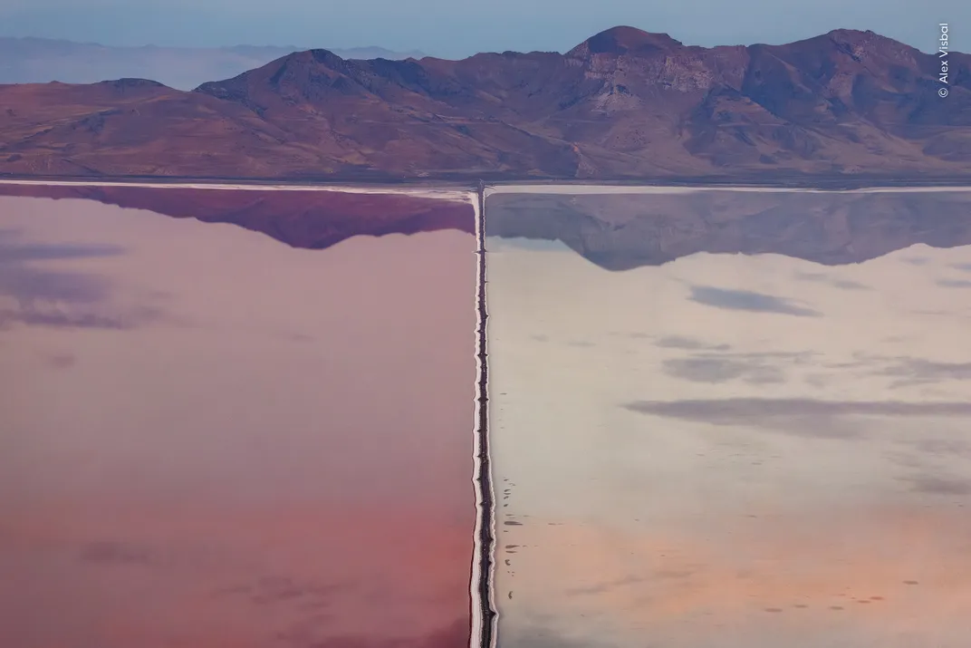 an overhead view of the Great Salt Lake cut through by a manmade barrier. On the left the water is pink, and on the right it is more clear. Mountains rise in the background