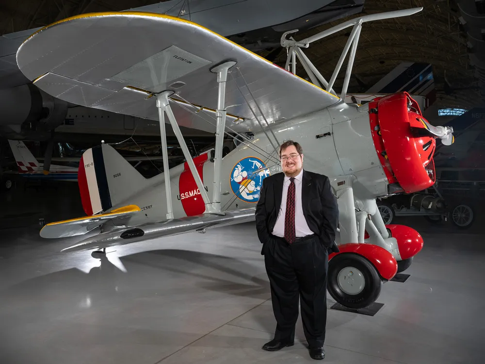 A white man in his 30s stands in front of a restored biplane displayed on a polished concrete floor inside a museum. He is wearing glasses and a dark suit with a white shirt and burgundy tie.