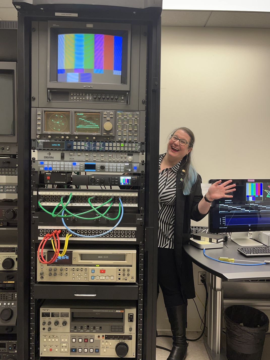 NMAAHC Video Archives Technician, Emily Nabasny smiles and waves to the camera from behind a towering rack of analog video equipment including a TV monitor displaying color bars.