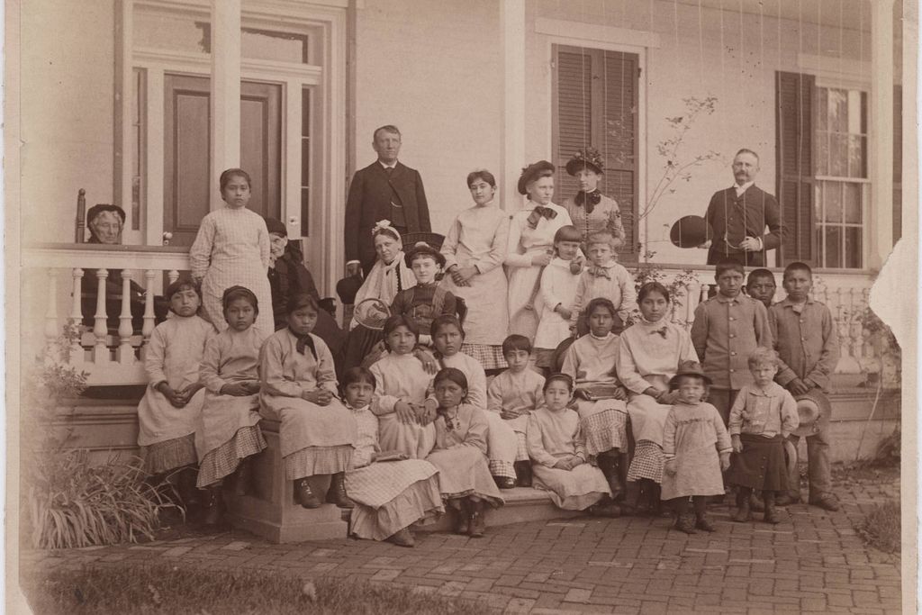 A sepia toned image of a group of young women, with a few young boys and girls, sitting in front of a white house on a porch. Pratt stands in dark garb behind the group