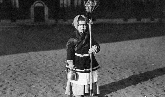 A young girl poses outside a building, wearing a dark coat, layers of skirts, and a kerchief over her hair, holding a broom in one hand and a kettle in the other. Black-and-white photo.
