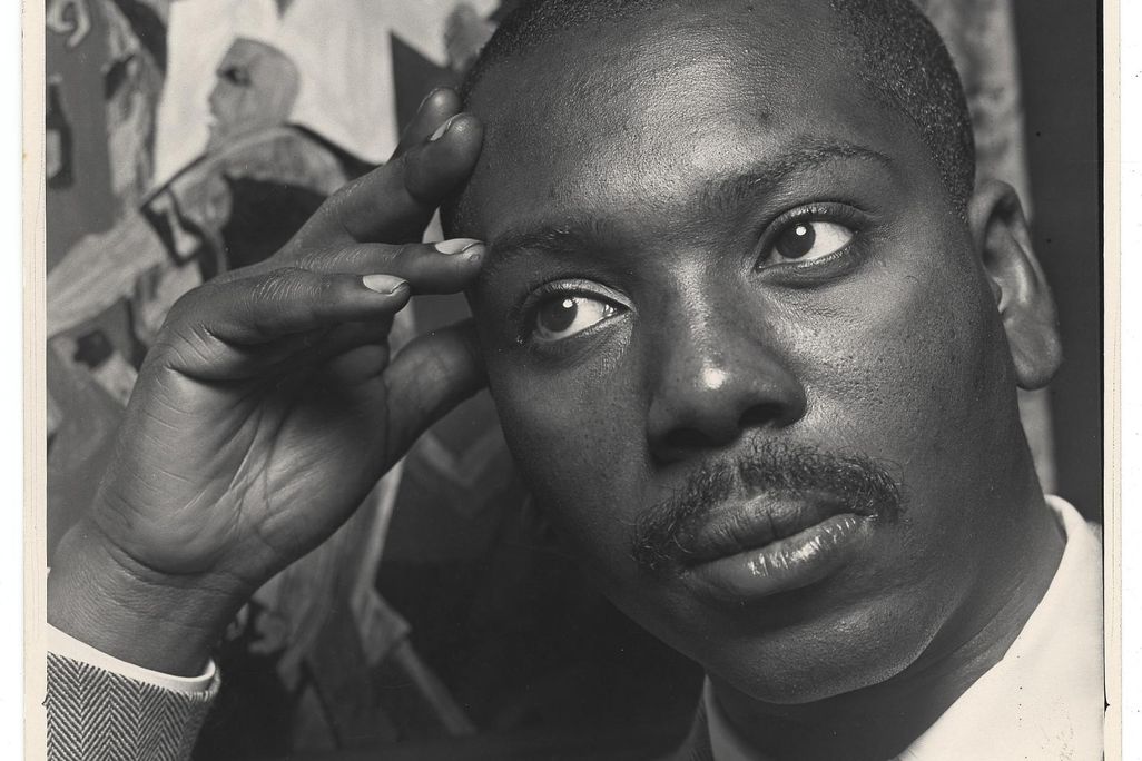 A black and white image of Lawrence, a black man with a mustache, looking contemplative with a suit and tie and resting his head against his hand, in front of one of his works of art