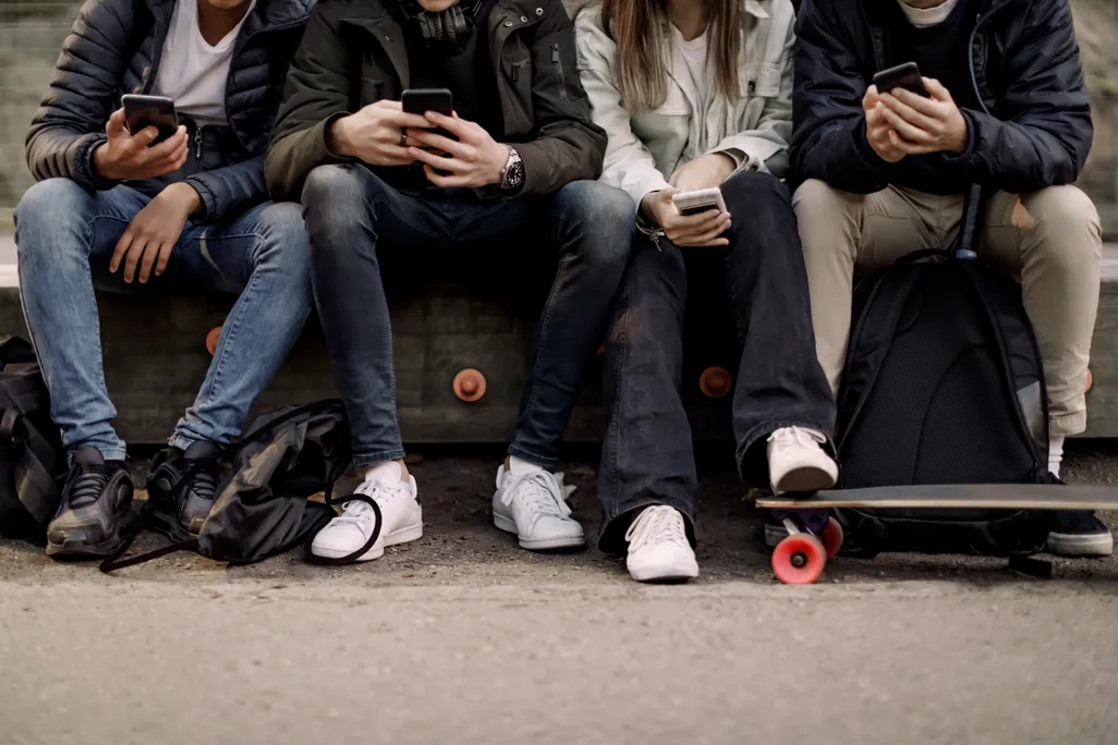 View from the waist down of kids sitting on a bench looking at their cellphones
