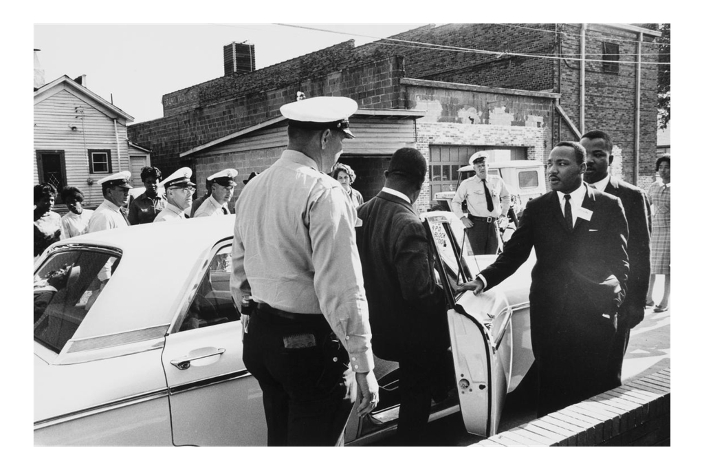 A black and white photograph of policeman in foreground, one man stepping into a squad car and MLK with an arm outstretched moving toward the car