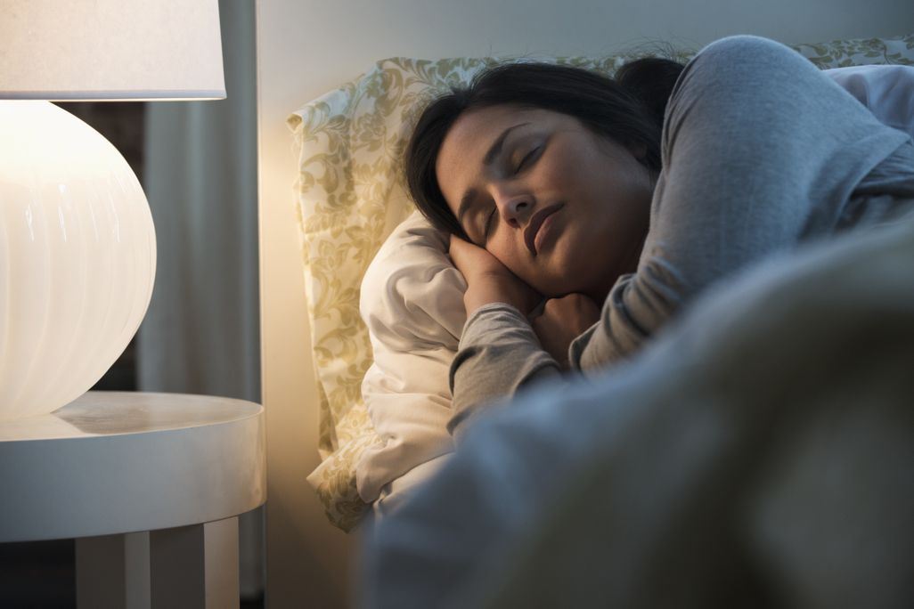 A woman sleeping in bed with a bedside lamp lit nearby
