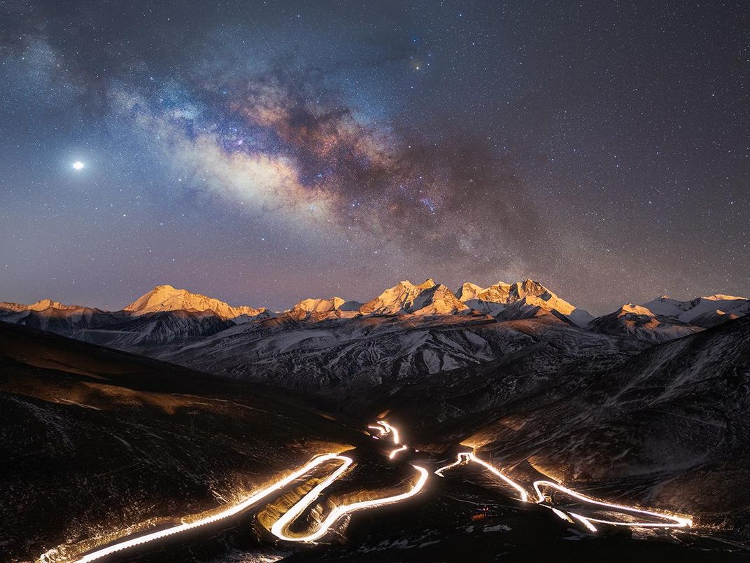 Una imagen de montañas contra el telón de fondo de la Vía Láctea.  Frente a la montaña hay una carretera iluminada por autos de alta velocidad.