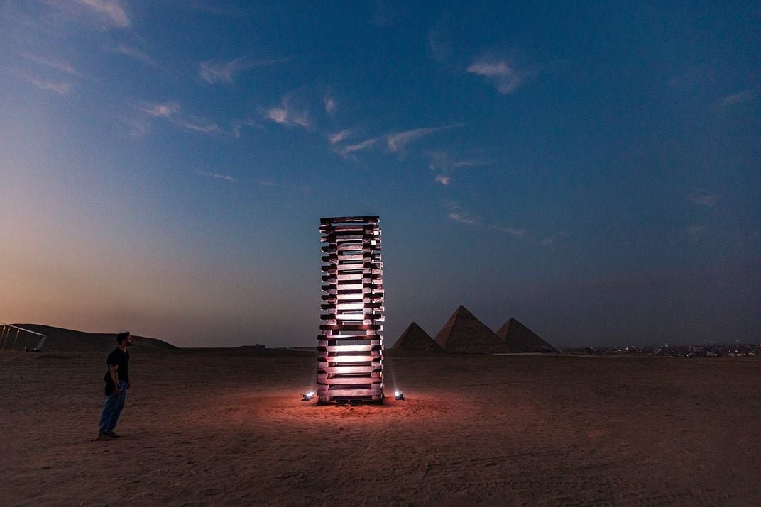 A stacked tower or ladder, lit up from within, stands in front of the three pyramids at dusk